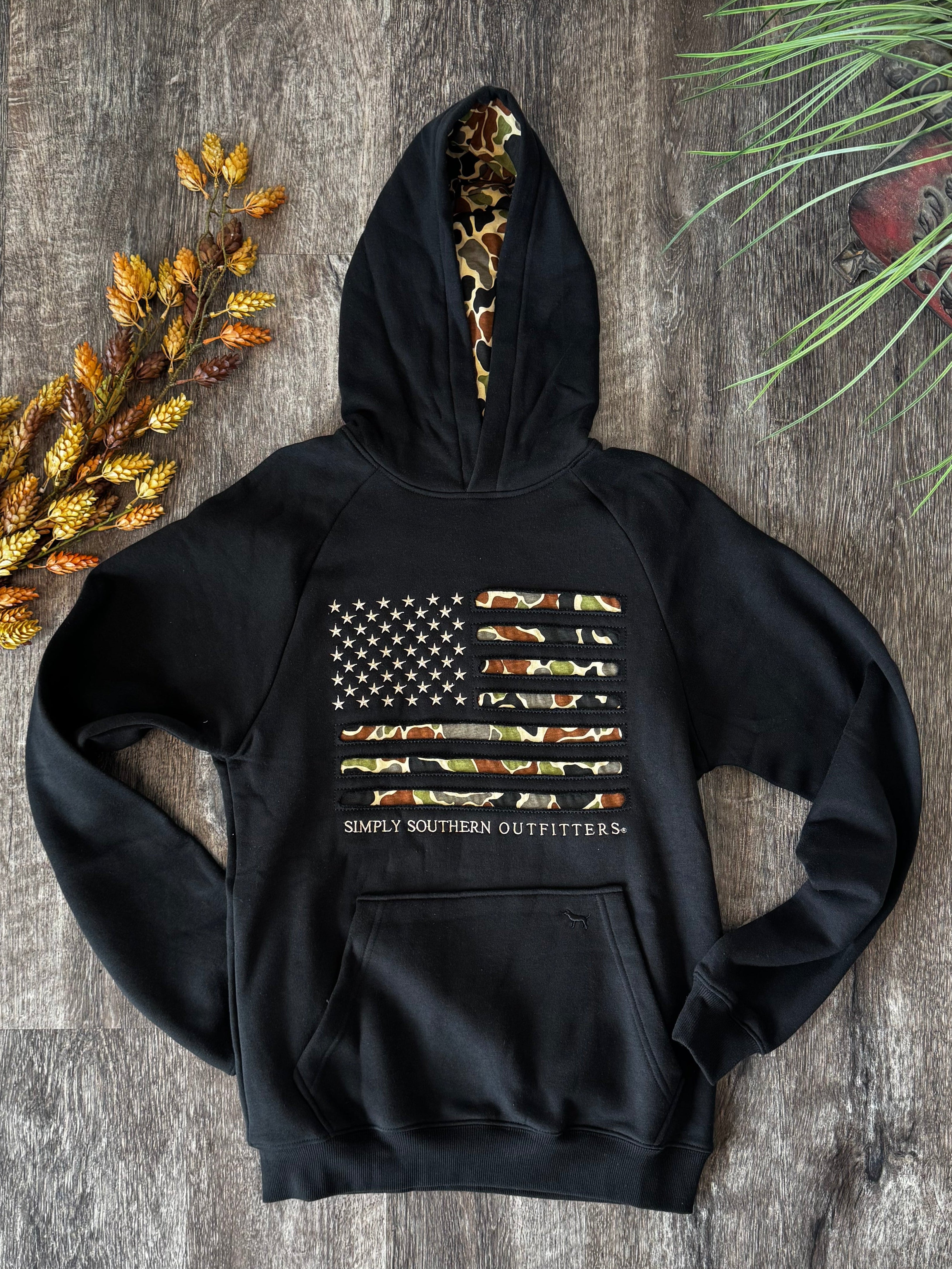 A black pullover hoodie laid flat on a gray wood floor, featuring a distressed American flag graphic on the chest made from camouflage-patterned strips, with the words 'SOUPY SOUTHERN OUTFITTERS' printed below. The hood lining shows a camouflage pattern, and the hoodie is styled with autumn leaves, a plant, and shoes at the bottom.